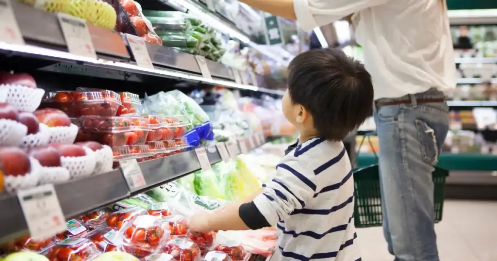 スーパーの青果コーナーで商品を選ぶ子どもと大人。親子が並んで野菜や果物を見ている様子。