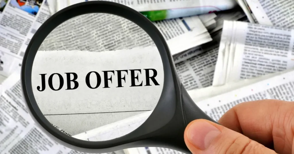 新聞の求人広告から『JOB OFFER』の文字を拡大鏡で覗く場面。ジョブ型採用の求人や採用理由を象徴するイメージ。