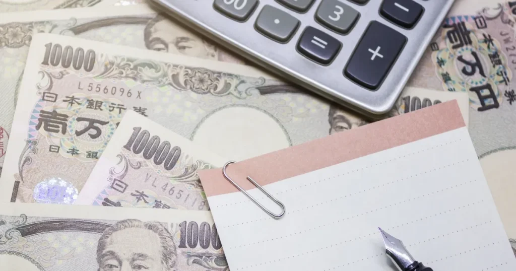 計算機と日本円の紙幣が並べられたテーブルに、メモ帳とペンが置かれている様子。転職活動の費用対策を考えるイメージ。