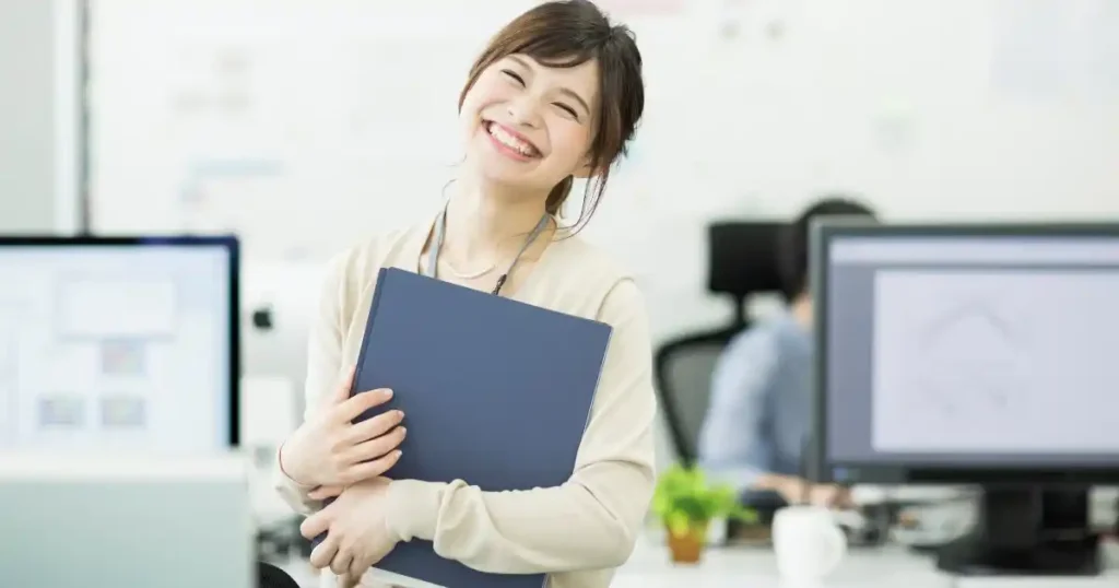 オフィス内でファイルを抱え笑顔を見せる女性。食品バイヤーに向いている人の特徴として、コミュニケーション力や責任感を象徴するイメージ。