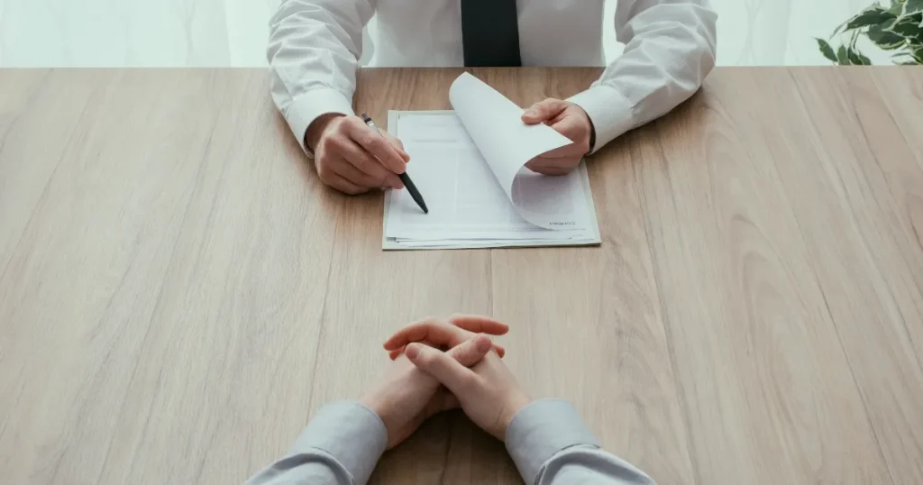 木目調のテーブルで資料を手に持ち、ペンを指し示すスーツ姿の人物と向き合う手元の様子。OBOG訪問の準備や持ち物を象徴するシーン。