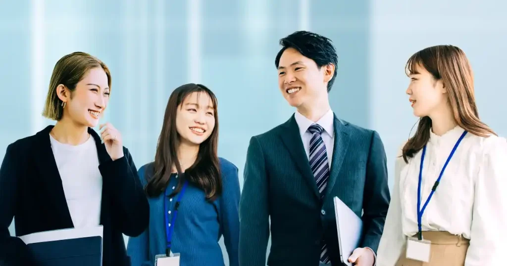 明るい表情で会話するスーツ姿の男性とビジネスカジュアルの女性たち。職場でのコミュニケーションやチームワークの重要性を象徴しています。
