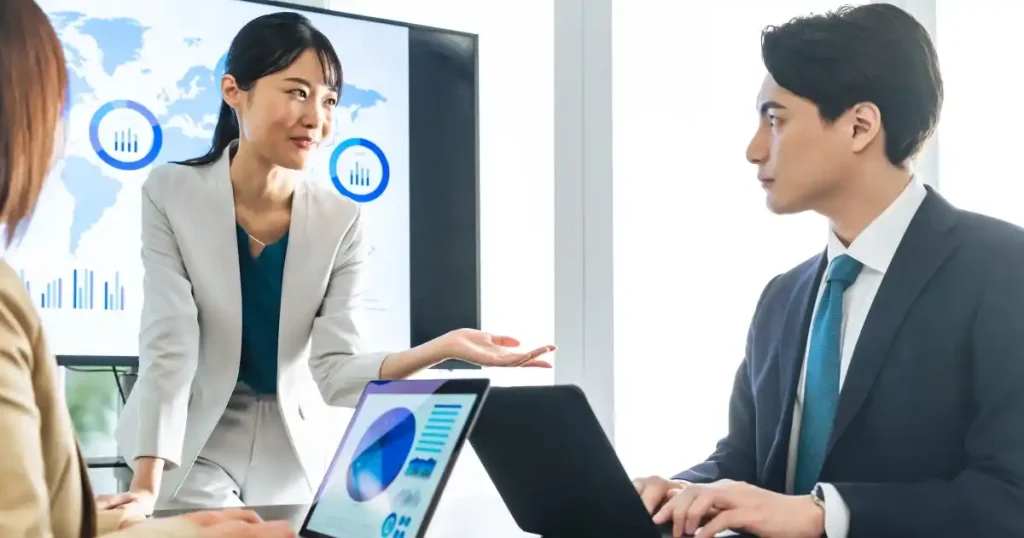 会議室でデータが表示された画面を背景に話し合うビジネスパーソンたち。マーケティング戦略の立案やキャリアパスの重要性を象徴する場面。
