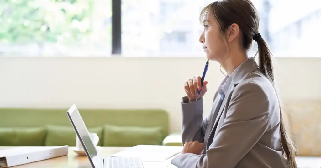 営業とコンサルの違いに関するQ&Aを説明するセクションの画像。スーツを着た女性がノートパソコンを前にペンを手に考え込んでいる様子。職業選択やキャリアについて検討している場面を象徴しています。