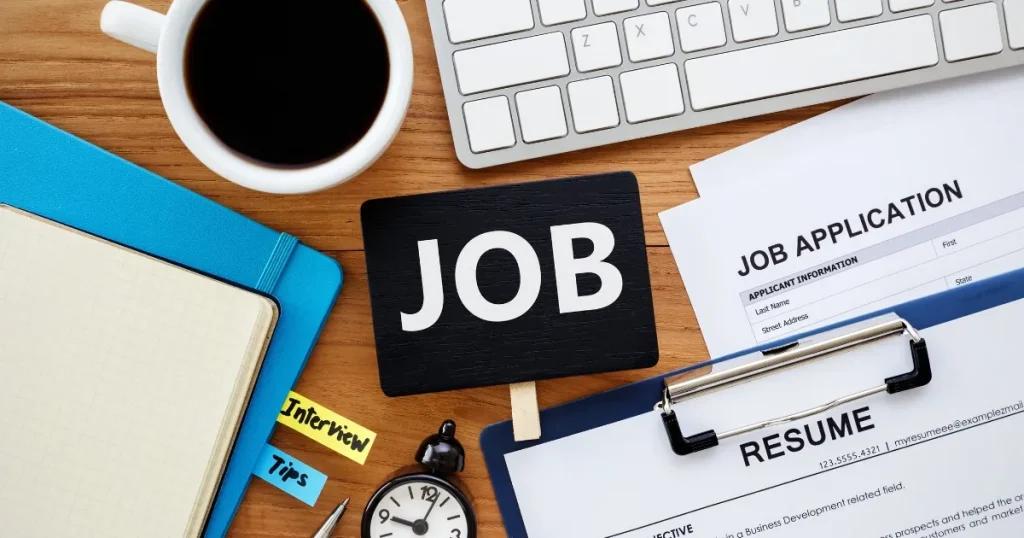 「JOB」の文字が書かれたプレート、履歴書、求人応募書類、ノート、コーヒーカップなどがデスクに並べられたシーン。求人票の重要性を象徴するイメージ。