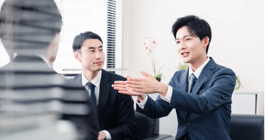営業とコンサルの役割を象徴する会議シーン。スーツ姿の男性が提案を説明し、他の参加者が話を聞いている場面。