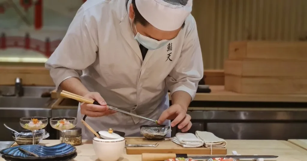 寿司職人が真剣な表情で魚を盛り付けている様子。カウンターには調理道具や器が並び、職人技が際立つシーン。東京で寿司職人を目指すための必要な準備やスキルを象徴しています。
