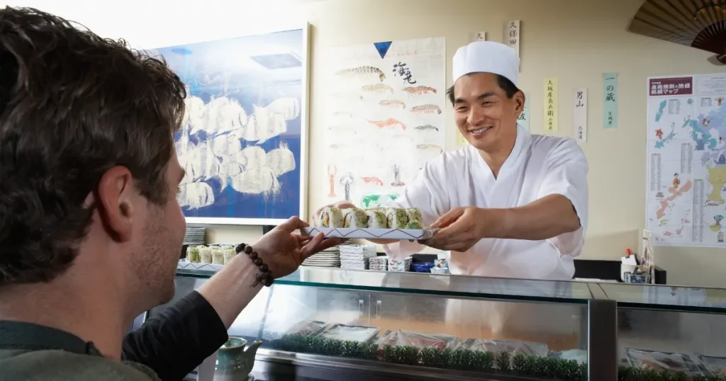 寿司職人がカウンター越しに客へ寿司を手渡すシーン。親しみやすい接客が、寿司職人業界での求人情報探しにおける人間関係の重要性を象徴している。