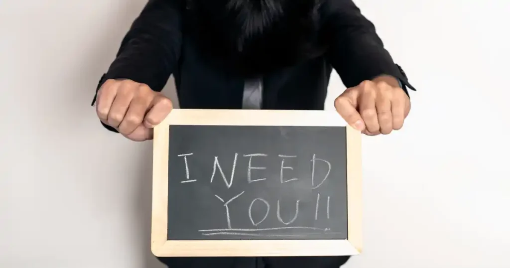 黒板に「I NEED YOU!!」と書かれたサインを持つ人物:人材不足や注目業界の求めるスキルを象徴するイメージ。