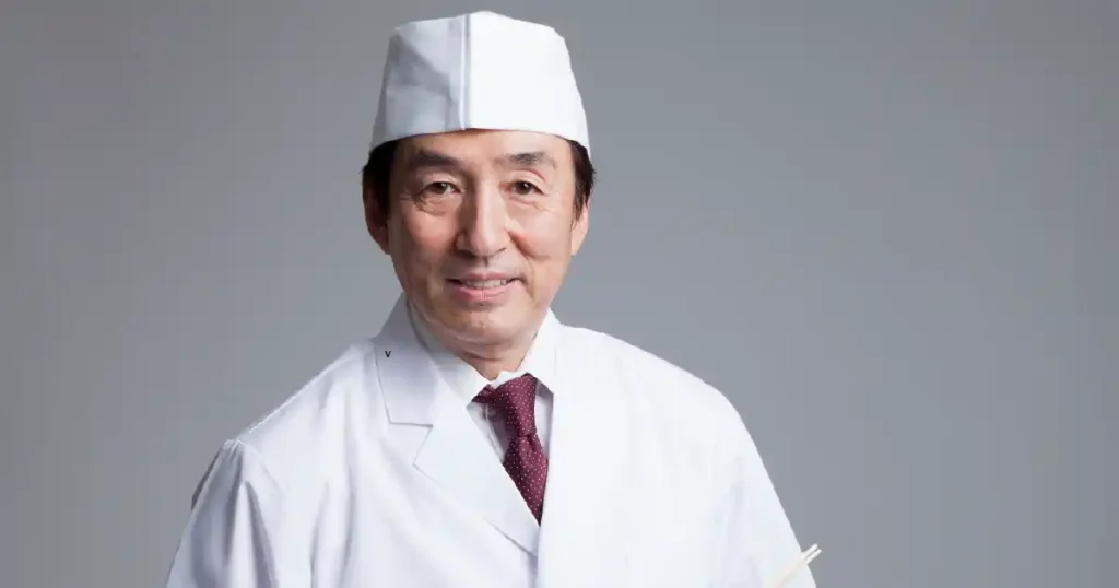 白衣と帽子を着用した寿司職人が微笑んでいる姿。職人の専門性と親しみやすさが伝わる。
