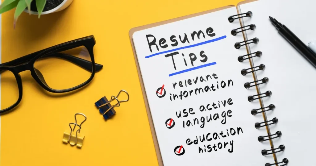 黄色の背景にノート、ペン、眼鏡、クリップが置かれ、ノートには「Resume Tips」として履歴書のポイントが記載されている：志望動機に関するNG行為を解説するセクションのイメージ。