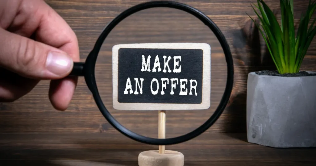 木製の背景に「MAKE AN OFFER」と書かれた小さな黒板が立てられ、虫眼鏡で拡大されている様子。横には観葉植物も置かれ、オファー型採用を象徴するビジュアル。