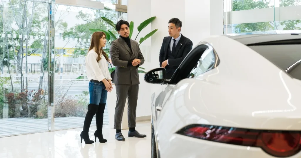 ディーラー店舗内で、新車について説明を受ける男女と営業スタッフ。明るいショールームにディスプレイされた白い車が特徴的で、ディーラーの役割を象徴する場面。