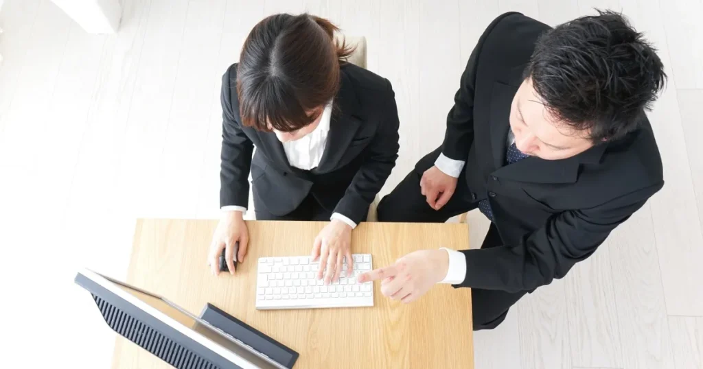 スーツを着た男女がデスクトップパソコンを使いながら協力して業務に取り組む様子。第2新卒としての転職のメリットを象徴する場面。