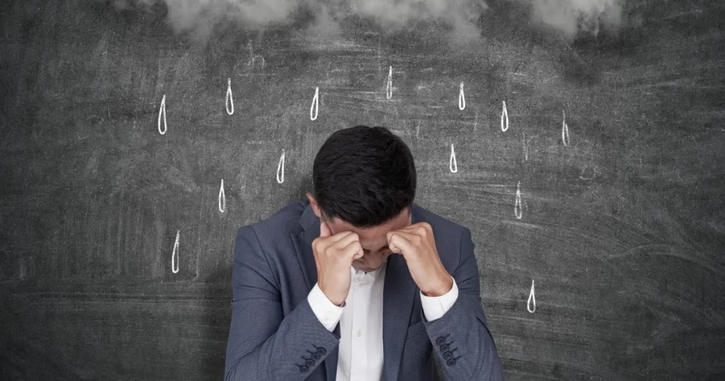 スーツを着た男性が頭を抱え、黒板に描かれた雨や雲が背景にあるイメージ。転職にまつわる不安や悩みを象徴するビジュアル。