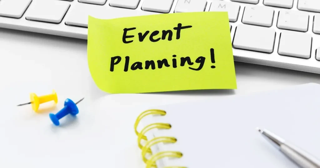キーボードの上に貼られた「Event Planning!」と書かれた黄色い付箋。手前にはノートやペン、カラフルな押しピンが配置され、イベントの計画を示唆するデザイン。