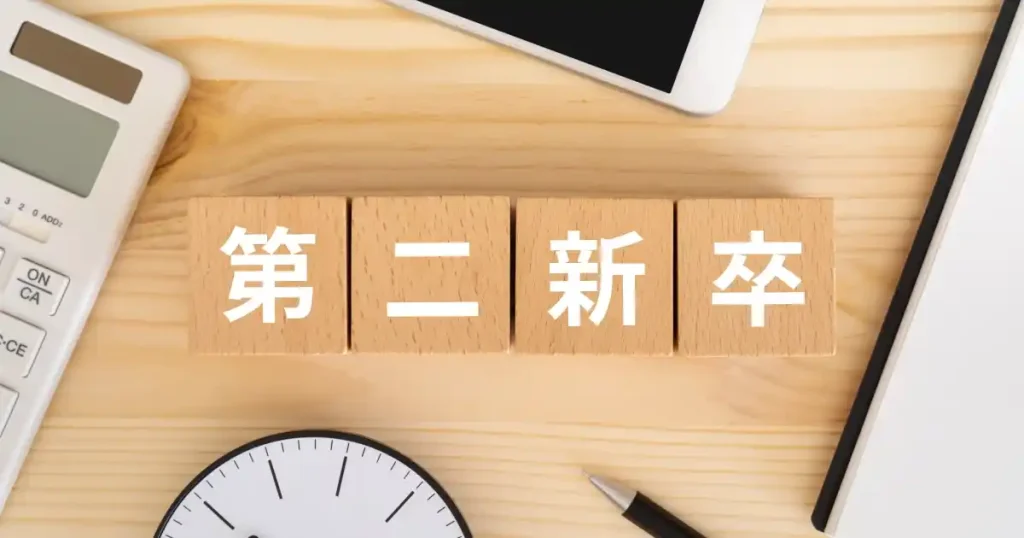 木製ブロックに「第二新卒」と書かれており、周囲には電卓、時計、スマートフォン、ペンが配置されているデスクのイメージ。第二新卒の定義や働き方を象徴するビジュアル。