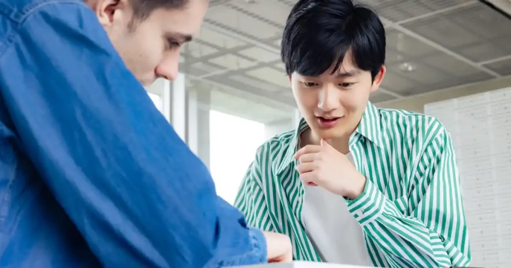 現代的なオフィスで話し合う二人の若者。片方が資料を見せながら説明し、もう一方が真剣に耳を傾けている様子が描かれている。