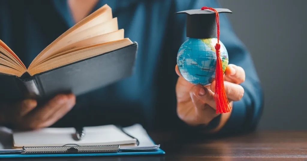 開いた本を手に持つ人物が、小さな地球儀に学位帽をかぶせたオブジェを掲げている。学びやグローバル経験を象徴するビジュアル。