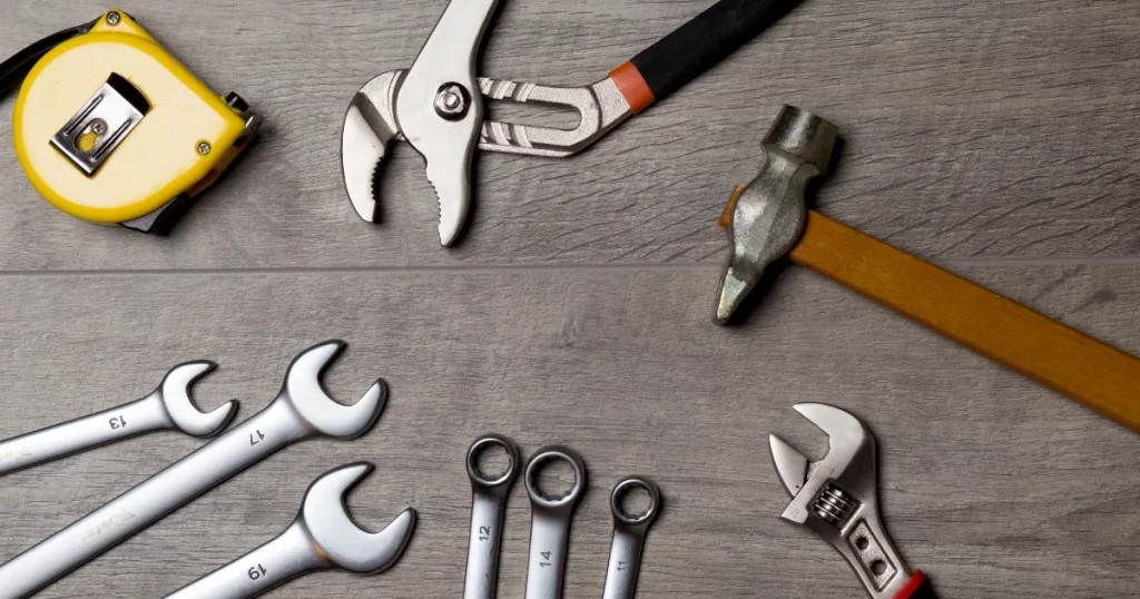 木目調の作業台に並べられた自動車整備に使用される工具が描かれた画像。スパナ、レンチ、ペンチ、ハンマー、メジャーなどの基本的な工具が含まれている。