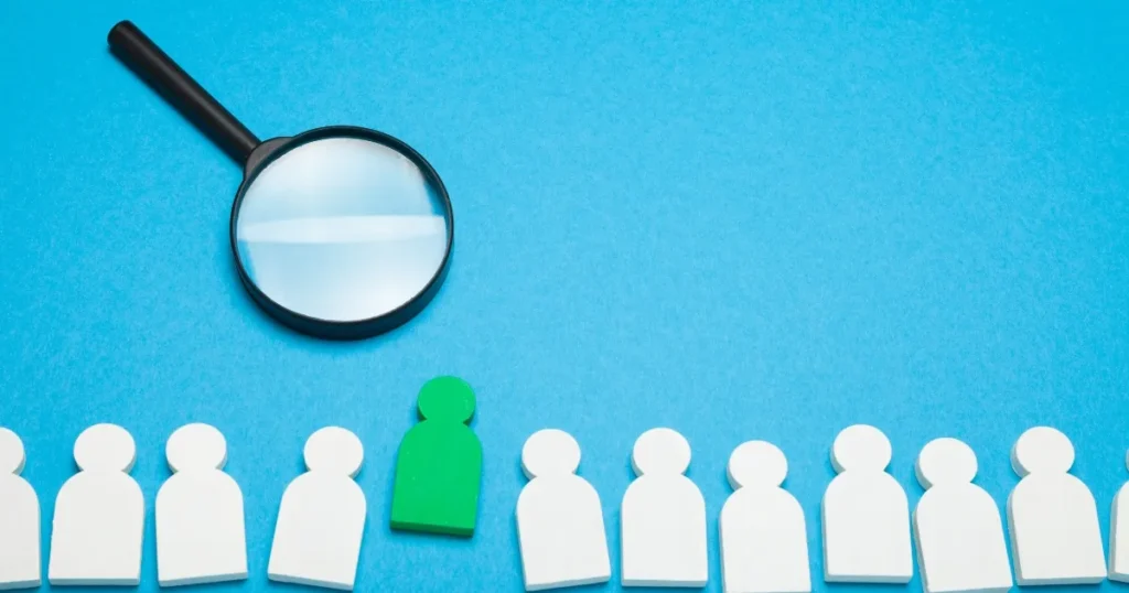 青い背景に並んだ白い人型のピースの中で、1つだけ緑色のピースが目立ち、上には拡大鏡が置かれている。ハイキャリア転職での候補者選定や戦略的アプローチを象徴するイメージ。