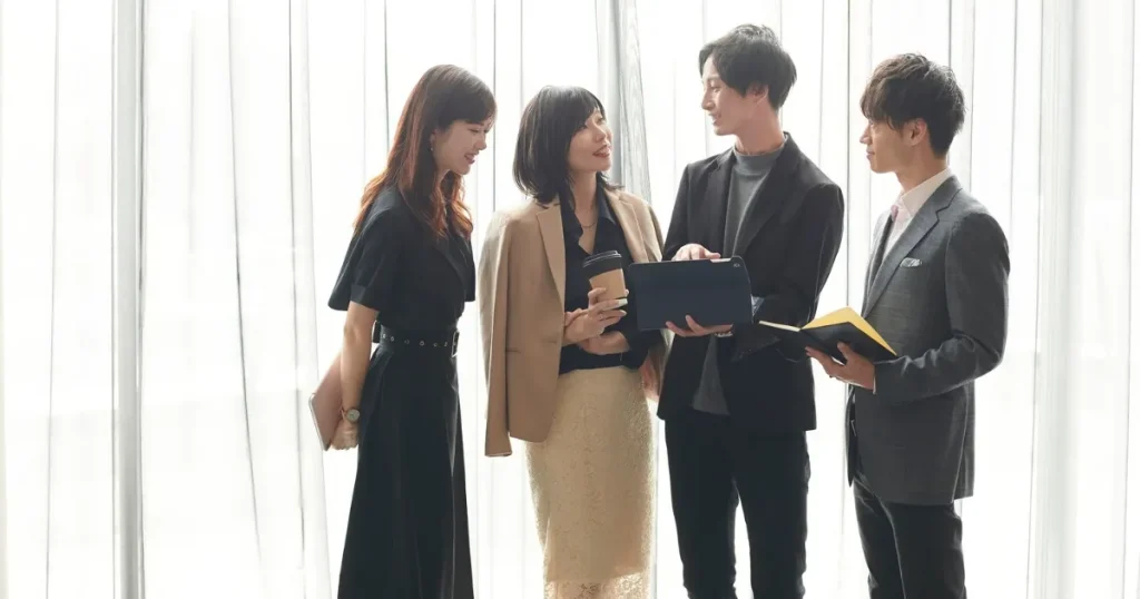 明るいオフィスで話し合う4人のビジネスパーソン。スーツやオフィスカジュアルを着用し、資料を手に持ちながら意見を交換している様子。