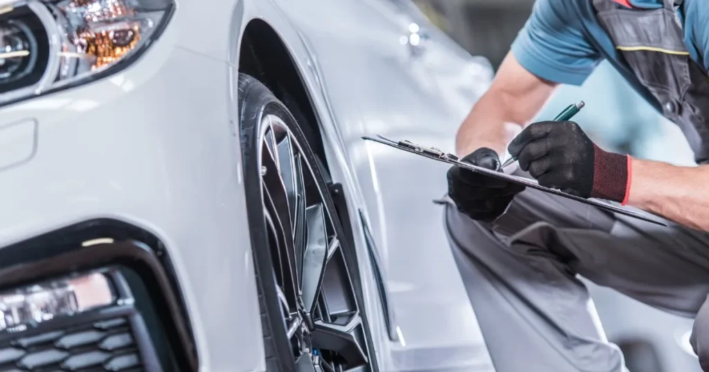 自動車整備士が白い車のタイヤ周辺を点検しながら、クリップボードに記録を書き込んでいる様子。