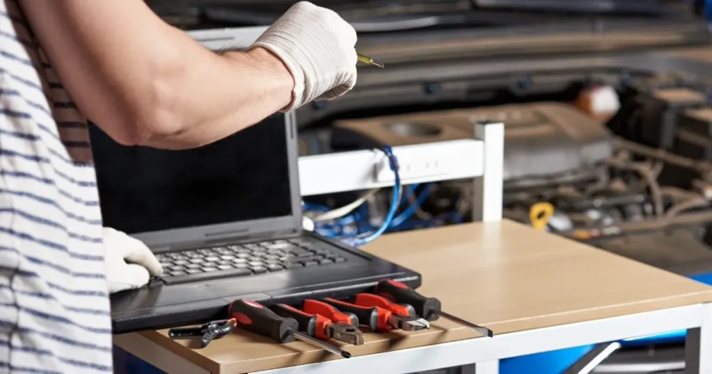 自動車整備士が手袋を着用し、整備用ツールとノートパソコンを使いながら作業している様子。背景には車両のエンジンルームが見える。
