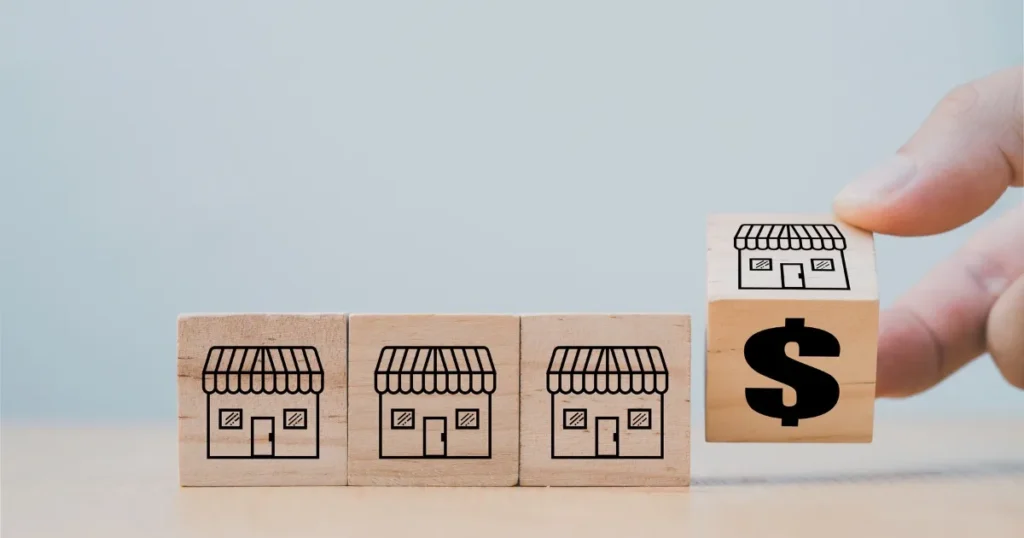 小さな店舗のイラストが描かれた木製ブロックが並び、その隣にドルマークが描かれたブロックを手で持ち上げている。スーパーの店舗開発職が収益性や成長に直結することを象徴するイメージ。