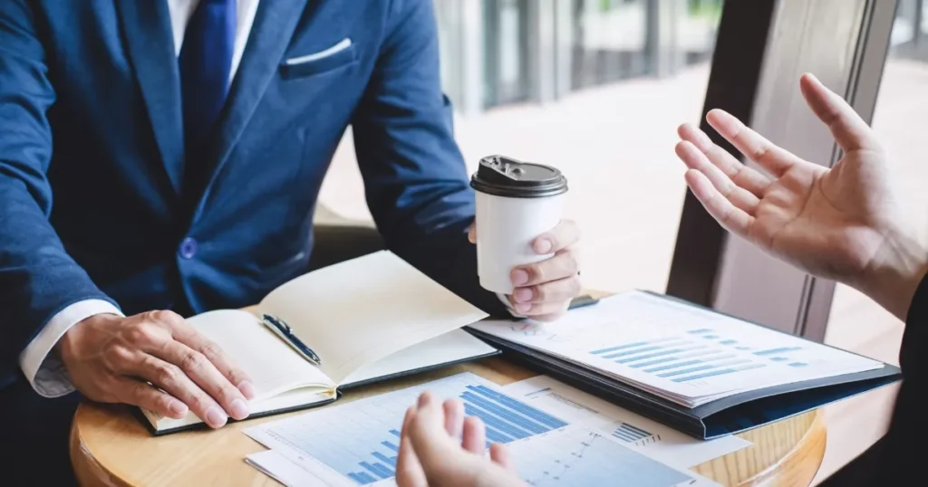 スーツ姿のビジネスマン2人がカフェで会話をしながら、テーブルに広げたグラフや資料を確認している。スーパーの店舗開発職における企画や戦略的な打ち合わせを象徴するイメージ。