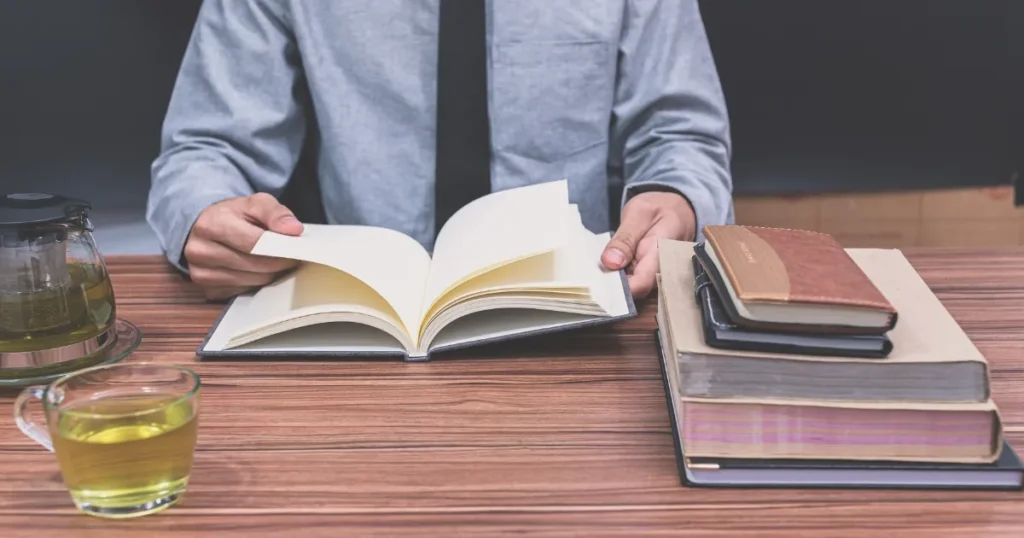 机の上に開いた本を読みながら、複数のノートや本が積まれている。隣にはお茶が置かれ、落ち着いた雰囲気で準備や学習に取り組む様子を表現している。