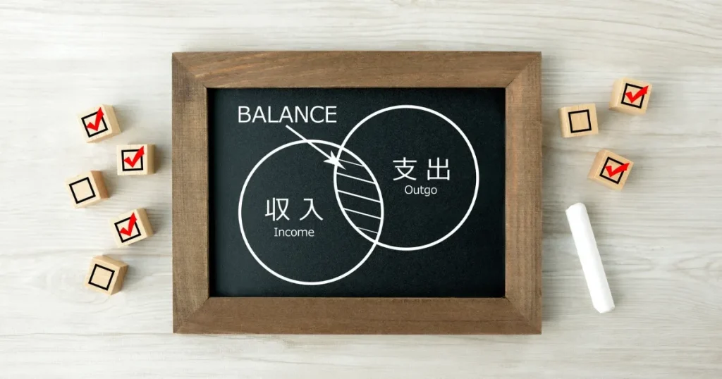 収入と支出のバランスを示す図が描かれた黒板とチェックマーク付きの積み木 転職による年収アップで注意すべきポイントを解説するセクションのイメージ画像