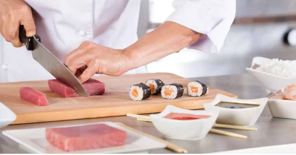 寿司職人が新鮮なマグロを丁寧に切り分ける様子。巻き寿司や調味料が並ぶ調理台で、寿司職人の現状を象徴するイメージ。