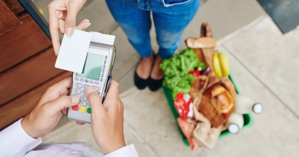 スーパーのレジでカード決済をする様子。買い物カゴには新鮮な食品がたくさん入っており、グロサリー部門の取扱商品を象徴するイメージ。
