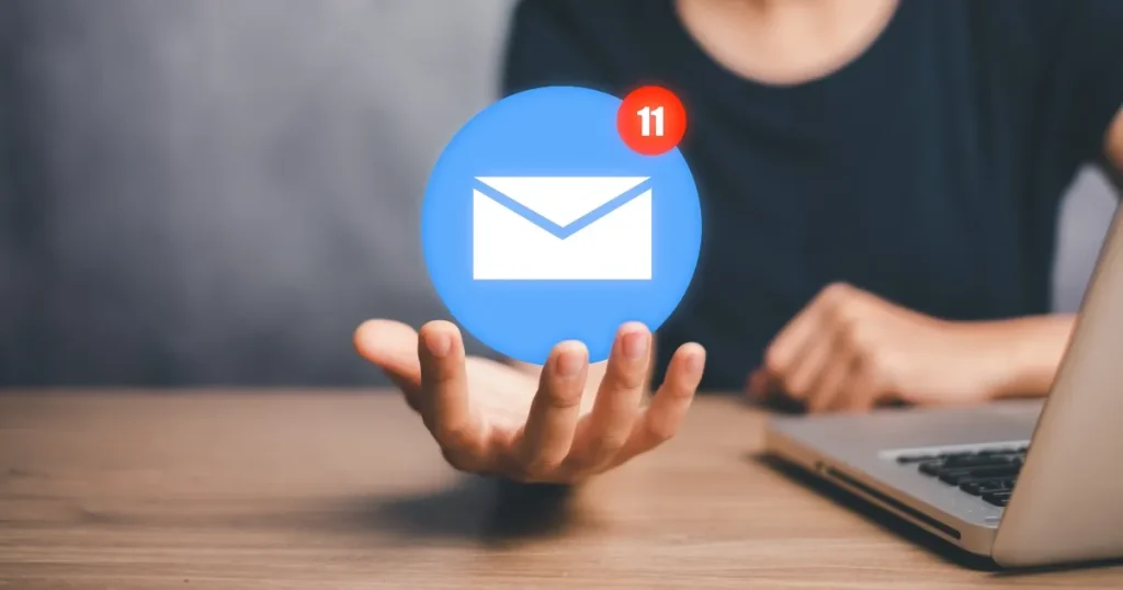 机の上で手を広げ、宙に浮かぶメール通知アイコンを支える人の姿。面接日程変更メールの具体的な例文を紹介するセクションに関連したビジュアル。