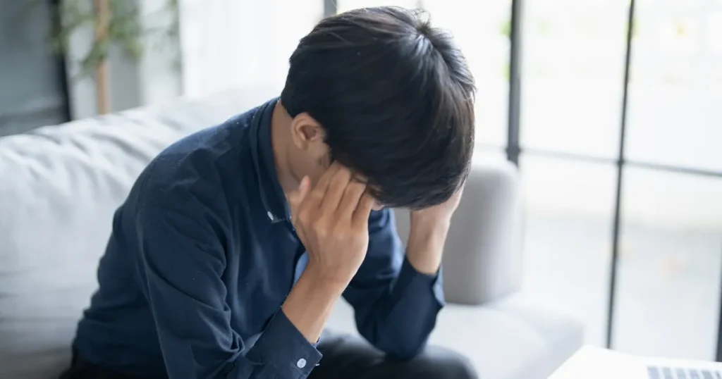 ソファに座って両手で顔を覆う男性。不動産営業のきついノルマや仕事の負担による悩みをイメージした写真。