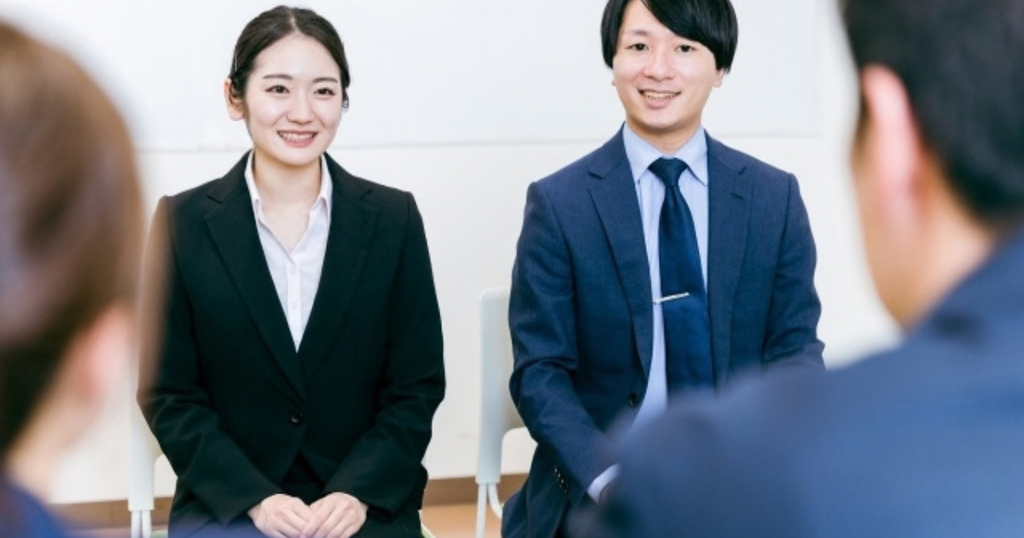 面接官の前で笑顔で座るリクルートスーツ姿の就活生2人で、特別な経験がなくてもガクチカは作れることをイメージした画像
